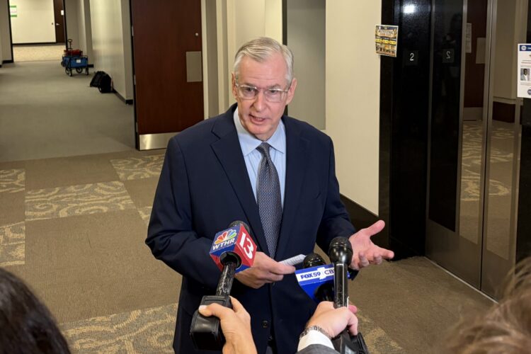 Greg Ballard stands in a hallway, taking questions from reporters holding microphones.