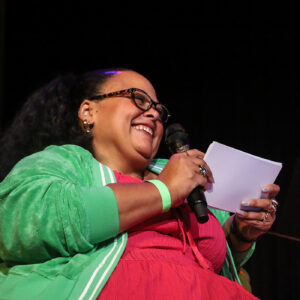 Pop of Culture cohost Kara DuQuette smiles while holding a microphone and note cards.
