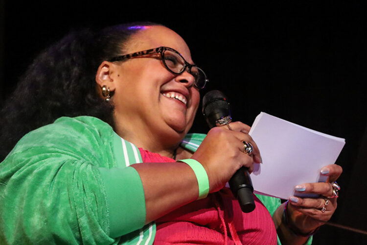 Pop of Culture cohost Kara DuQuette smiles while holding a microphone and note cards.