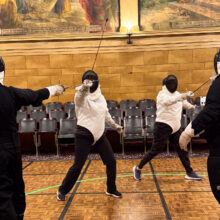 Two people clad in padded white protective gear and helmets spar with fencing rapiers against opponents dressed in black.