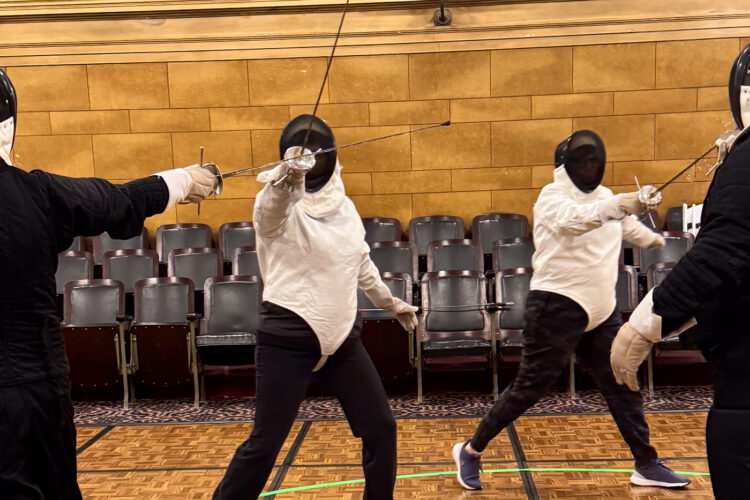 Two people clad in padded white protective gear and helmets spar with fencing rapiers against opponents dressed in black.