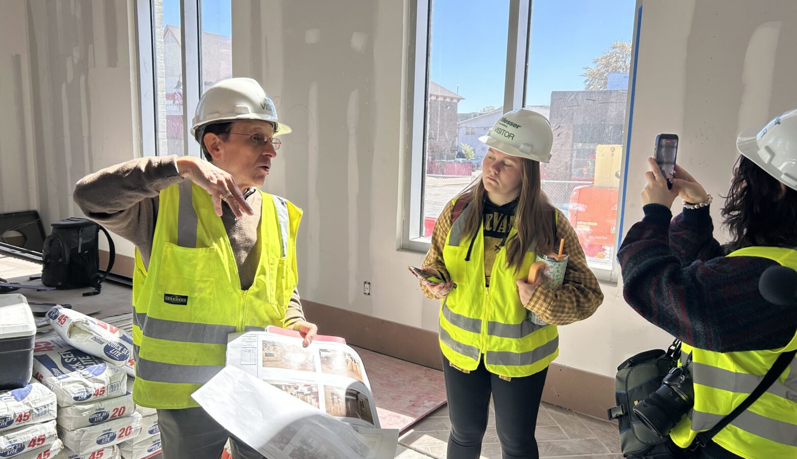 A man in a high visibility vest holds papers depicting room layouts. He talks to a group of people. They all stand inside of an unfinished room in a building.