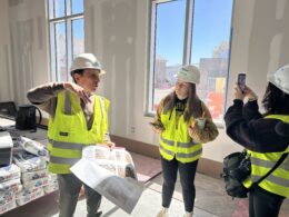 A man in a high visibility vest holds papers depicting room layouts. He talks to a group of people. They all stand inside of an unfinished room in a building.