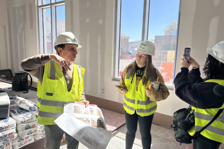A man in a high visibility vest holds papers depicting room layouts. He talks to a group of people. They all stand inside of an unfinished room in a building.
