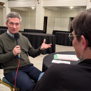 Kerry Glann talks with Jen Blackmer in the middle of an empty banquet hall. He is holding a microphone and gesticulating.