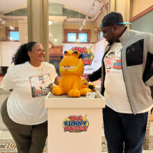 Kara DuQuette and Deon Parson pose on either side of a podium with a statuette of Garfield the cat. The podium and Deon's t-shirt read "The Funny Pages," the name of the exhibition they are standing in.Kara DuQuette and Deon Parson pose on either side of a podium with a statuette of Garfield the cat. The podium and Deon's t-shirt read "The Funny Pages," the name of the exhibition they are standing in.