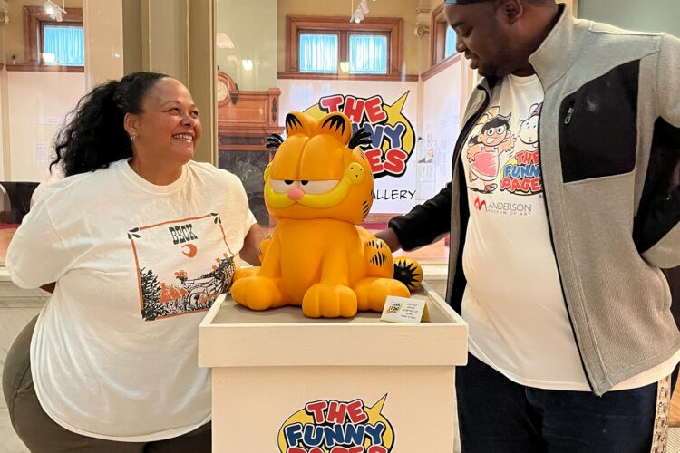Kara DuQuette and Deon Parson pose on either side of a podium with a statuette of Garfield the cat. The podium and Deon's t-shirt read "The Funny Pages," the name of the exhibition they are standing in.Kara DuQuette and Deon Parson pose on either side of a podium with a statuette of Garfield the cat. The podium and Deon's t-shirt read "The Funny Pages," the name of the exhibition they are standing in.