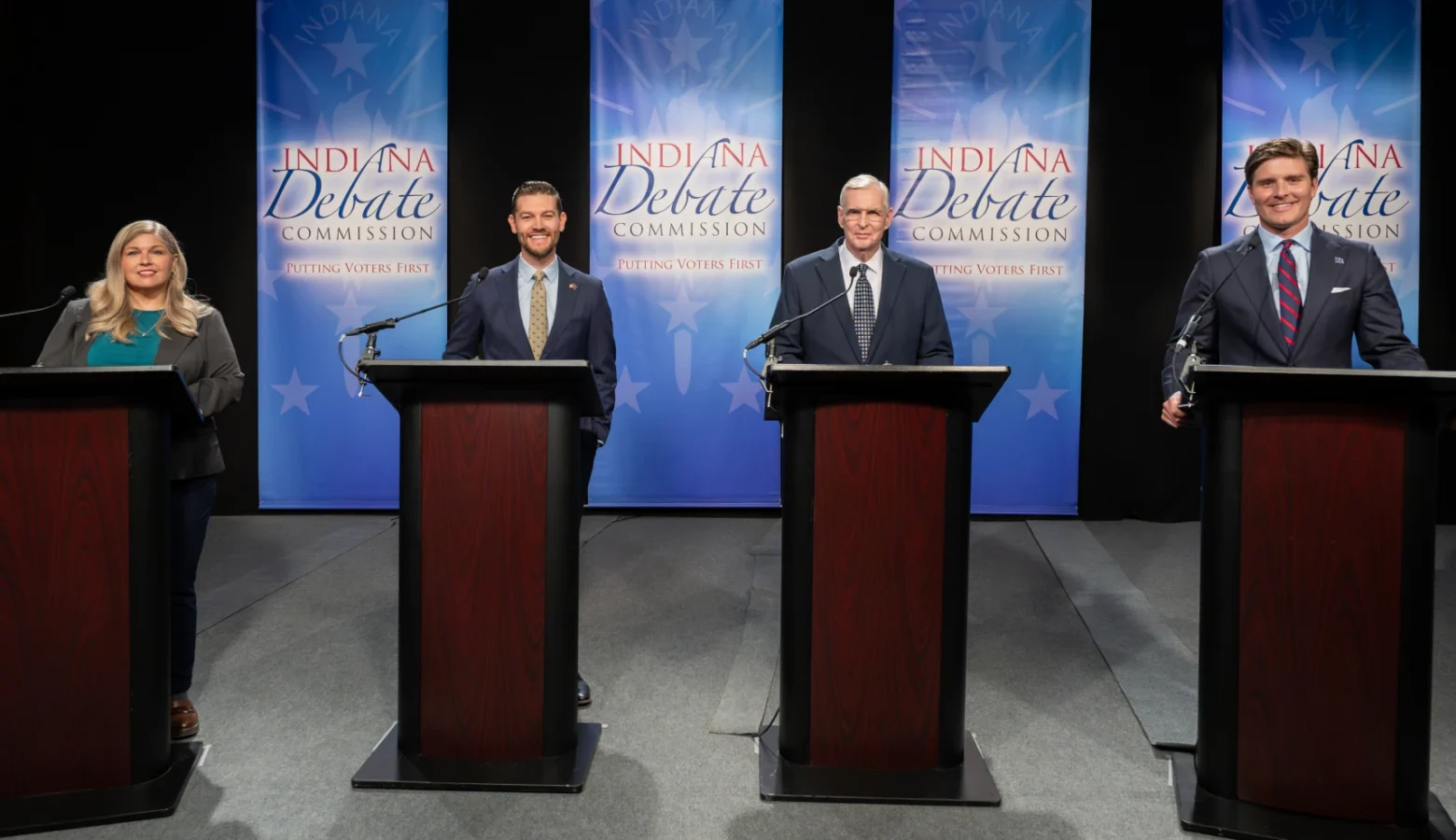 Indiana Secretary of State candidates gathered Wednesday for a debate held by the Indiana Debate Commission at WFYI studios. The candidates are standing behind dark wooden podiums in front of a light blue background that says "Indiana Debate Commission" on four individual separated by black curtains.