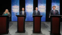 Indiana Secretary of State candidates gathered Wednesday for a debate held by the Indiana Debate Commission at WFYI studios. The candidates are standing behind dark wooden podiums in front of a light blue background that says "Indiana Debate Commission" on four individual separated by black curtains.