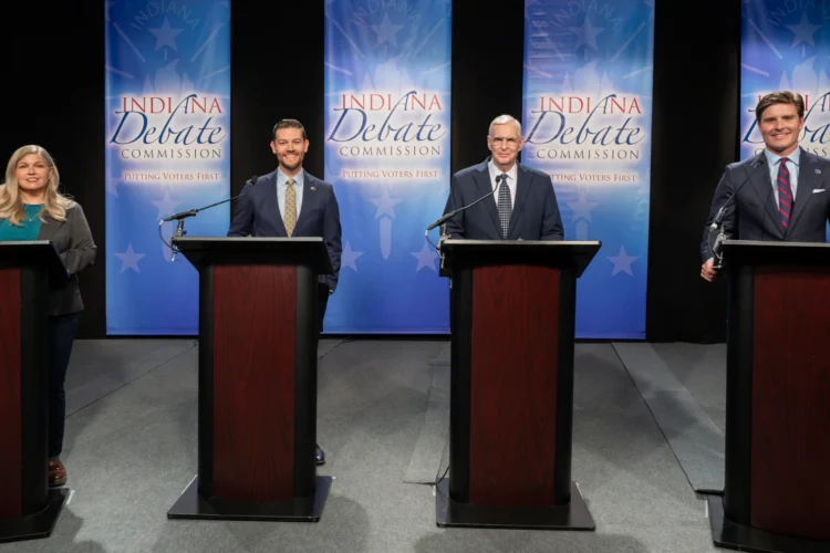 Indiana Secretary of State candidates gathered Wednesday for a debate held by the Indiana Debate Commission at WFYI studios. The candidates are standing behind dark wooden podiums in front of a light blue background that says "Indiana Debate Commission" on four individual separated by black curtains.