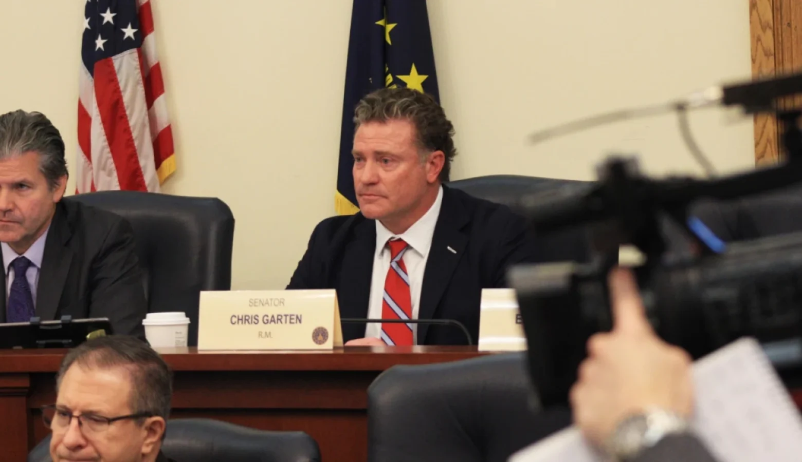 Sen. Chris Garten (R-Charlestown) sparred with democrats over the effectiveness of tax abatements to businesses. He is seated at a committee table in a statehouse conference room with American and Indiana flags behind him and other committee members seated on either side of him. A TV news camera is in the foregroud.