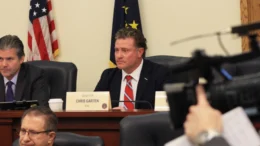 Sen. Chris Garten (R-Charlestown) sparred with democrats over the effectiveness of tax abatements to businesses. He is seated at a committee table in a statehouse conference room with American and Indiana flags behind him and other committee members seated on either side of him. A TV news camera is in the foregroud.