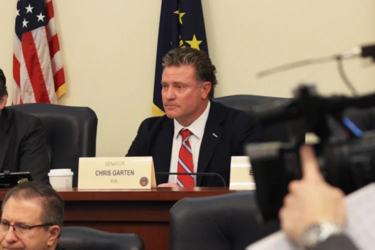 Sen. Chris Garten (R-Charlestown) sparred with democrats over the effectiveness of tax abatements to businesses. He is seated at a committee table in a statehouse conference room with American and Indiana flags behind him and other committee members seated on either side of him. A TV news camera is in the foregroud.