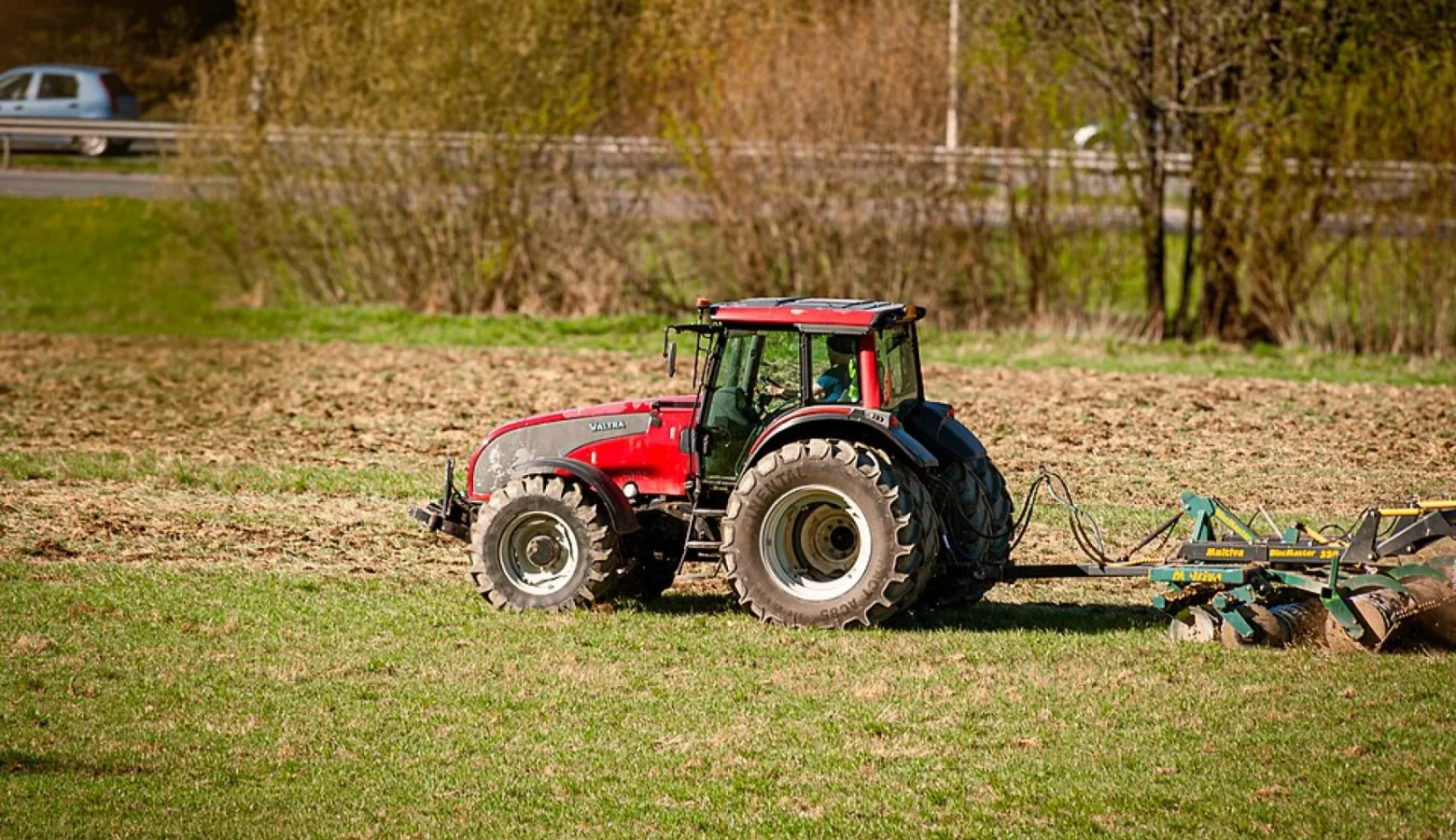 A large red tractor is pulling a discing attachment on a field that appears to have grass in prep for being turned over. Small trees are on the far boundary of the field.
