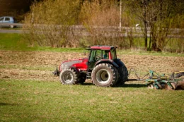 A large red tractor is pulling a discing attachment on a field that appears to have grass in prep for being turned over. Small trees are on the far boundary of the field.