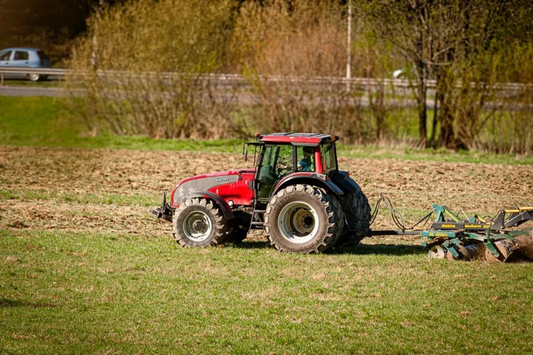 A large red tractor is pulling a discing attachment on a field that appears to have grass in prep for being turned over. Small trees are on the far boundary of the field.