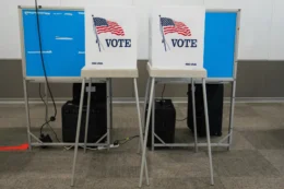 The photo shows two voting booths, side by side with a white partition on each displaying an American flag rippling in the wind and the word "VOTE" in large capital letters.