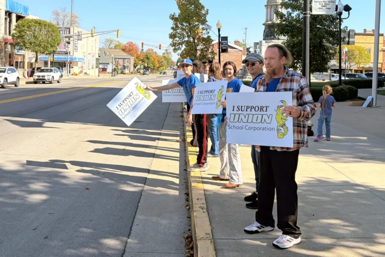 Protesters stand along a road, carrying signs that say I Support Union School Corporation.